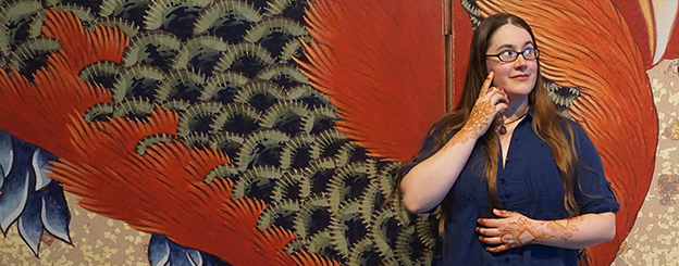 Portrait of the artist smiling in front of a wall with artwork of a gigantic pheonix.